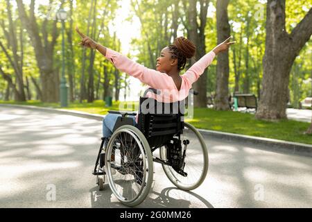 Donna nera portatrice di handicap in sedia a rotelle a piedi al parco cittadino, che mostra un gesto di vittoria, sentirsi positiva e motivata Foto Stock