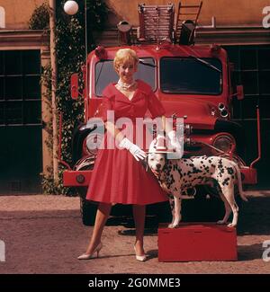 Alla moda negli anni '50. Una giovane donna in un abito rosso, guanti bianchi e scarpe bianche è in piedi di fronte a un camion da fuoco Ford insieme a un cane dalmata con un casco capo fuoco americano. Il dalmatier è tradizionalmente una mascotte della brigata del fuoco. Svezia 1958 rif CV68-2 Foto Stock