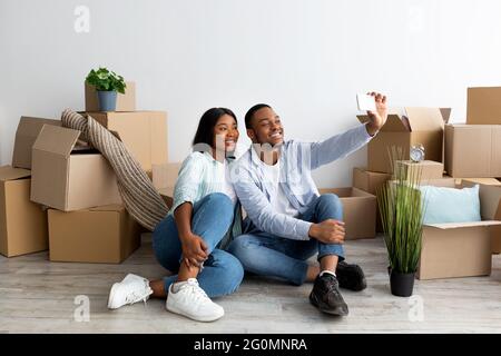 Felice coppia nera che si sposta a casa propria e prendendo selfie tra scatole di cartone, spazio libero Foto Stock