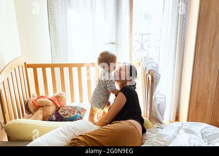 La mamma sorridente si trova sul letto vicino al bambino in piedi sullo sfondo di una culla e giocattoli Foto Stock