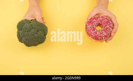 Confronto delle abitudini alimentari. Donna che tiene broccoli e ciambella su sfondo giallo. Spazio di cimasa. Foto Stock