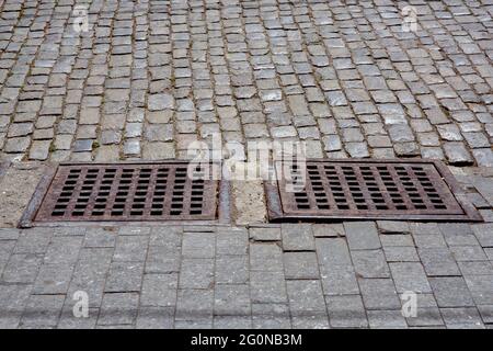 sistema di drenaggio delle infrastrutture della città vecchia con grata di ferro arrugginito di boccata di sistema di acque piovane di marciapiede da piastrelle di granito vicino alla strada da pavimentazione Foto Stock