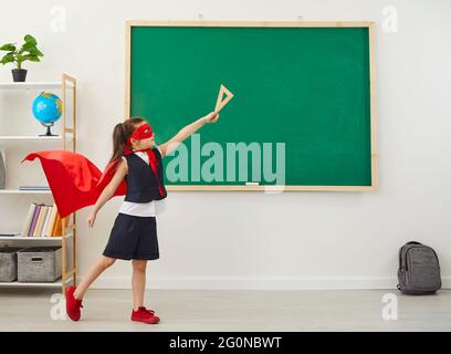 Ragazza carina in costume super eroe che posa vicino a lavagna in classe. Foto Stock