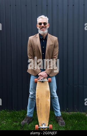 intera lunghezza di felice uomo di mezza età in occhiali da sole che tengono la longboard all'esterno Foto Stock