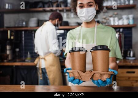 Tazze di carta nelle mani del barista afroamericano in maschera medica su sfondo sfocato Foto Stock