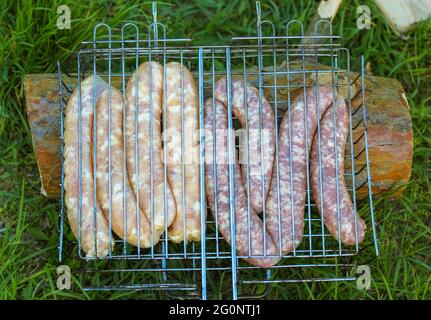 Salsiccia cruda su una griglia barbecue top vista dall'alto, piatto, preparazione di carne fresca all'aperto. Picnic nel bosco in una giornata estiva soleggiata. Sfondo verde erba. Foto Stock