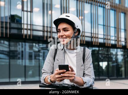 Donna sorridente appoggiata su uno scooter elettrico a spinta utilizzando un telefono cellulare mentre si trova vicino a un edificio di uffici Foto Stock