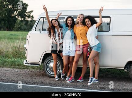 Felici amici diversi al camper van. Quattro donne sorridenti in piedi insieme sulla strada vicino a una macchina durante la vacanza. Foto Stock