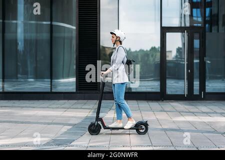 Vista laterale di una giovane donna d'affari che guida uno scooter elettrico a spinta. Femmina in bicicletta casco guida scooter elettrico contro l'edificio. Foto Stock