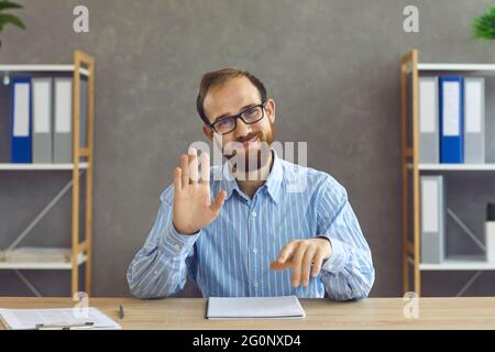 Felice lavoratore in ufficio in occhiali seduti alla scrivania, sorridendo e muovendo la mano alla macchina fotografica Foto Stock