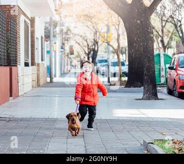 Felice ragazzino che corre con il suo cane dachshund su un marciapiede della città. Foto Stock