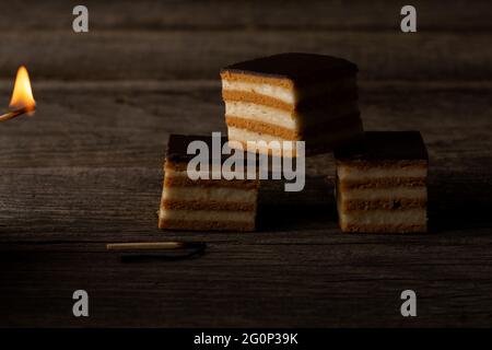 Tre fette di torta di Gerbeaud, illuminate dal fuoco di un fiammifero ardente, giacciono su tavole di legno intemperie. Foto Stock