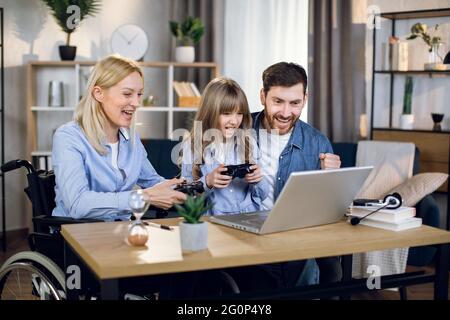 Donna disabile che gioca video giochi a casa con la sua bella figlia e bel marito. Genitori e bambini che usano un computer portatile moderno e joystick per l'intrattenimento. Foto Stock