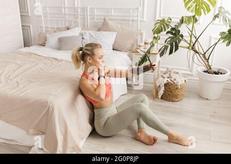 Bella istruttore di yoga femminile utilizzando smartphone, facendo l'allenamento online da casa, sorridendo. Felice giovane donna in elegante sportswear che fa un selfie Foto Stock