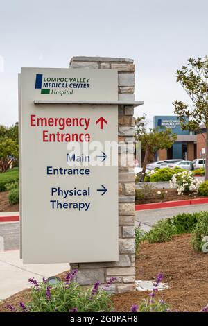 Lompoc, CA, USA - 26 maggio 2021: Emblema bianco su blu sul cartello di direzione all'ingresso della strada del Lompoc Valley Medical Center. Auto in parcheggio e impianti Foto Stock