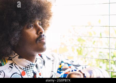 closeup di giovane ragazzo nero con capelli afro e baffi in profilo accanto a una finestra con griglia in metallo sfocato Foto Stock