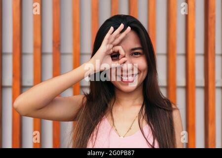 Attraente giovane donna che fa il segno di destra e che copre il suo occhio, esprime emozione, modella pone all'aperto Foto Stock