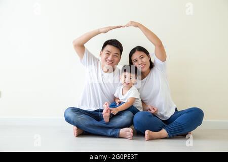 Papà e mamma che fanno casa di figura del tetto con le braccia delle mani sopra le teste, Famiglia asiatica con il figlio seduto sul pavimento nella stanza, casa residenziale di nuova costruzione pur Foto Stock