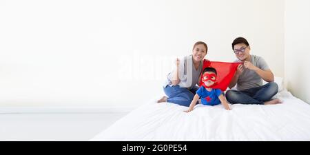 Padre, madre e figlio asiatici stanno giocando il supereroe sul letto in camera da letto. La famiglia è cordiale e si diverte Foto Stock