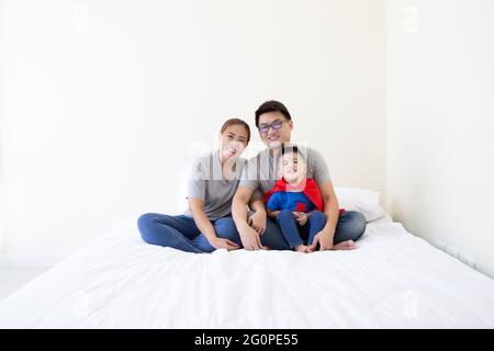 Padre, madre e figlio asiatici stanno giocando il supereroe sul letto in camera da letto. La famiglia è cordiale e si diverte Foto Stock