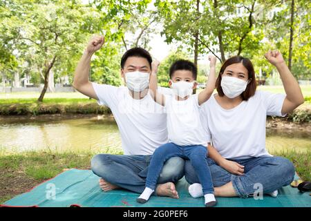 Famiglia asiatica indossare maschera medica di protezione per prevenire il virus Covid-19 e mano su e seduta insieme sul pavimento al verde giardino pubblico. Famiglia prote Foto Stock