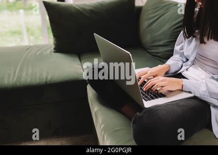 Una bambina di brunette si siede su un divano verde e stampa o sta digitando qualcosa su un computer portatile Foto Stock