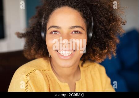 Primo piano ritratto di allegra e ottimista giovane donna dalla pelle scura con un paio di cuffie ricci afro, naturale bellezza mista donna in cuffia con microfono guarda la fotocamera Foto Stock