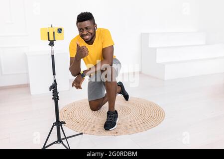 Bel giovane maschio è seduto di fronte alla fotocamera dello smartphone, facendo una lezione video sullo stile di vita sportivo. Afro American fitness Trainer dà Foto Stock