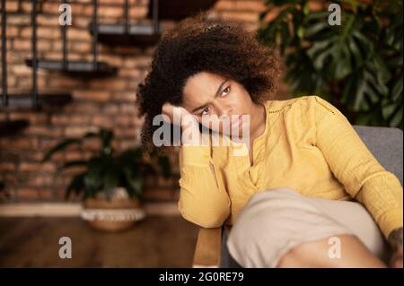 Concetto di depressione. La donna afro-americana sconvolta si sente annoiata seduto sul divano a casa da sola, la donna nera disperata si sente sola, manca il ragazzo, donna multirazziale preoccupata per il futuro Foto Stock