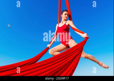 acrobat atletico, gymnast aggraziato che esegue l'esercizio aereo con tessuti rossi all'aperto su sfondo cielo. donna flessibile in tuta rossa esegue circo Foto Stock