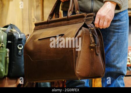 La mano di un uomo che tiene una borsa da viaggio marrone sullo sfondo di altre borse nel negozio. Il concetto di prodotti in pelle naturale Foto Stock
