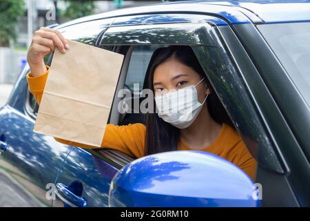 Donna asiatica indossando maschera e tenendo sacchetto di carta di fast food attraverso la finestra auto. Concetto di servizio di ristorazione drive-through Foto Stock