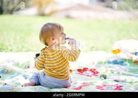 Il bambino si siede su una coperta colorata sull'erba verde, tiene un cetriolo in mano e gnaws un vaso di plastica. Vista posteriore Foto Stock