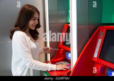 Giovane donna asiatica che ritira denaro con una carta alla macchina automatica, femmina in piedi presso ATM della banca Foto Stock