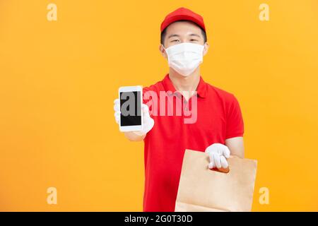 Felice giovane asiatico consegna uomo in uniforme rossa, maschera medica viso, guanti di protezione portare borsa mostrando telefono cellulare Foto Stock