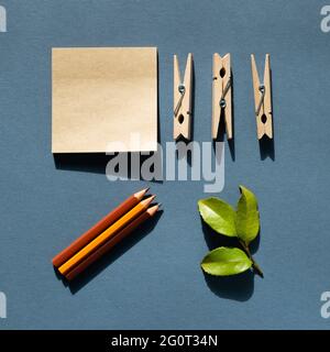Blocco note e matite colorate e clip in legno e foglia verde sulla scrivania. Sfondo blu marino. Luogo di lavoro e di studio. Disposizione piatta, vista dall'alto, spazio di copia Foto Stock