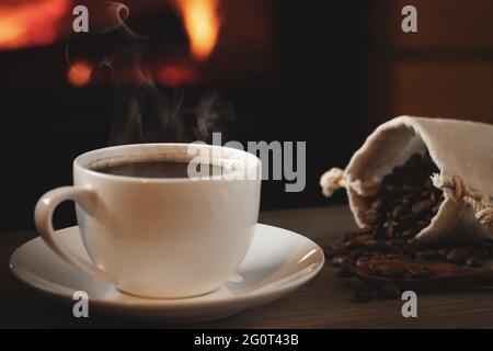 Primo piano di una tazza di caffè caldo e di chicchi di caffè in una borsa su un tavolo di legno di fronte a un caminetto acceso. Messa a fuoco selettiva. Foto Stock