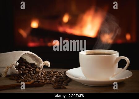 Tazza di caffè caldo e chicchi di caffè in una borsa su un tavolo di legno di fronte a un camino acceso. Messa a fuoco selettiva. Foto Stock