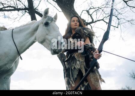 Donna in immagine dell'antico guerriero amazzone spara un arco e mira a bersaglio vicino al suo cavallo tra la foresta. Foto Stock