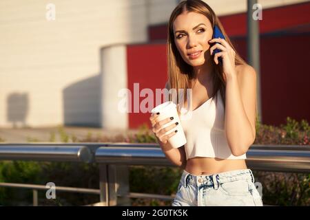 Giovane donna in attesa del treno cittadino e che parla al telefono con il caffè in mano. Il passeggero in tram ferma le chiamate con lo smartphone. Concetto di trasporto pubblico e di comunicazione. Foto di alta qualità Foto Stock