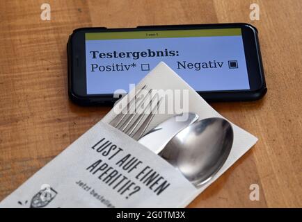 03 giugno 2021, Brandeburgo, Potsdam: Un tovagliolo con forchetta, coltello e cucchiaio e l'impronta 'Fancy an aperitivo?' Si trova in un ristorante accanto a uno smartphone il cui display mostra un risultato negativo del test Corona. Le nuove regole Corona per la gastronomia del Brandeburgo hanno portato a fastidio in molti stabilimenti. Questo perché, a partire da giovedì, il requisito di test per i clienti nei ristoranti all'aperto viene rinunciato solo se l'ospite non offre posti a sedere interni. In vista del clima soleggiato, molti stabilimenti hanno quindi deciso, secondo un sondaggio del dpa, di offrire inizialmente solo posti a sedere sulle terrazze. Foto Stock