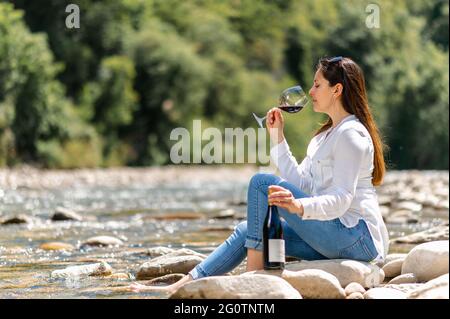 Bella donna che beve vino rosso seduto su una roccia sul lungofiume. Foto Stock