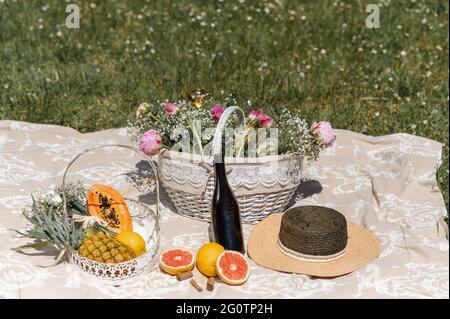 Coperta da picnic su un prato con cesto pieno di fiori, frutta tropicale, un cappello di paglia e una bottiglia di vino. Foto Stock