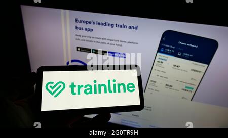 Persona in possesso di smartphone con logo della piattaforma di biglietti britannica trainline plc sullo schermo di fronte alla pagina web dell'azienda. Mettere a fuoco il display del telefono. Foto Stock