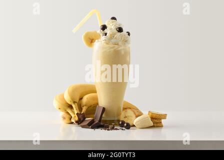 Frullato di banana con cioccolato e crema decorato con porzioni di frutta e cioccolato intorno su tavolo di legno bianco e sfondo isolato chiaro. Dal Foto Stock