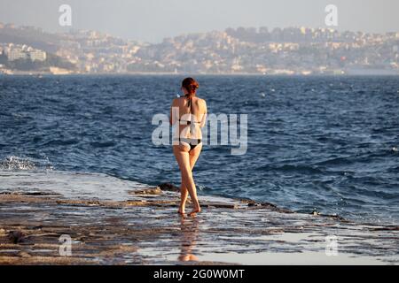 Vacanza in spiaggia, donna in bikini a piedi con smartphone sulla costa di pietra sul mare blu e sullo sfondo della città Foto Stock