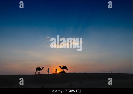 Silhouette di due cammelli e i loro cammelli alle dune di sabbia del deserto di Thar, Rajasthan, India. Nuvola con il sole che tramonta, cielo sullo sfondo. Cameleers Foto Stock