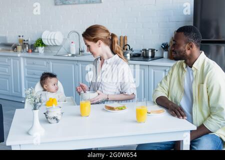 madre che tiene la ciotola vicino alla bambina e al marito afroamericano durante la colazione Foto Stock