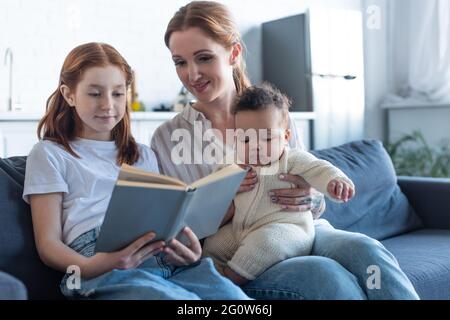 ragazza sorridente che legge libro vicino a madre e sorella africana americana infantile Foto Stock