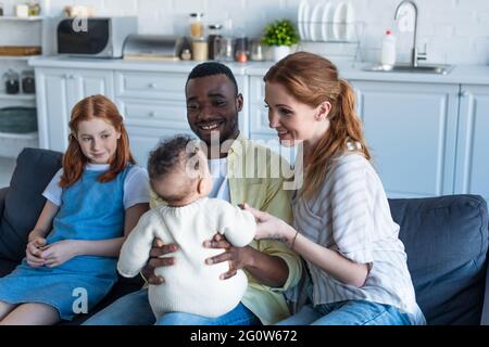 felice uomo afroamericano che tiene il bambino infante vicino alla famiglia multietnica sorridente Foto Stock